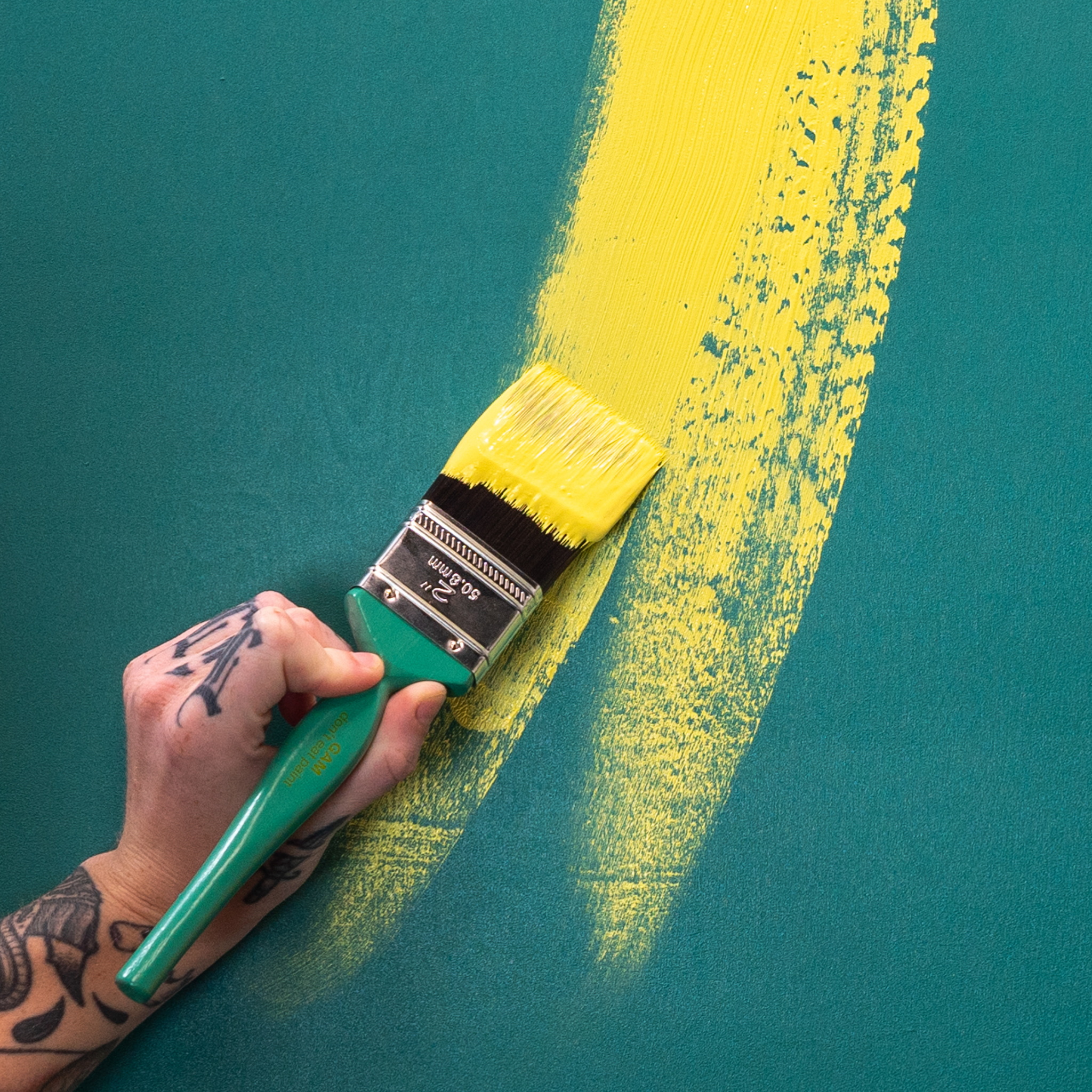 A tattooed artist's hand painting with yellow using a large paint brush