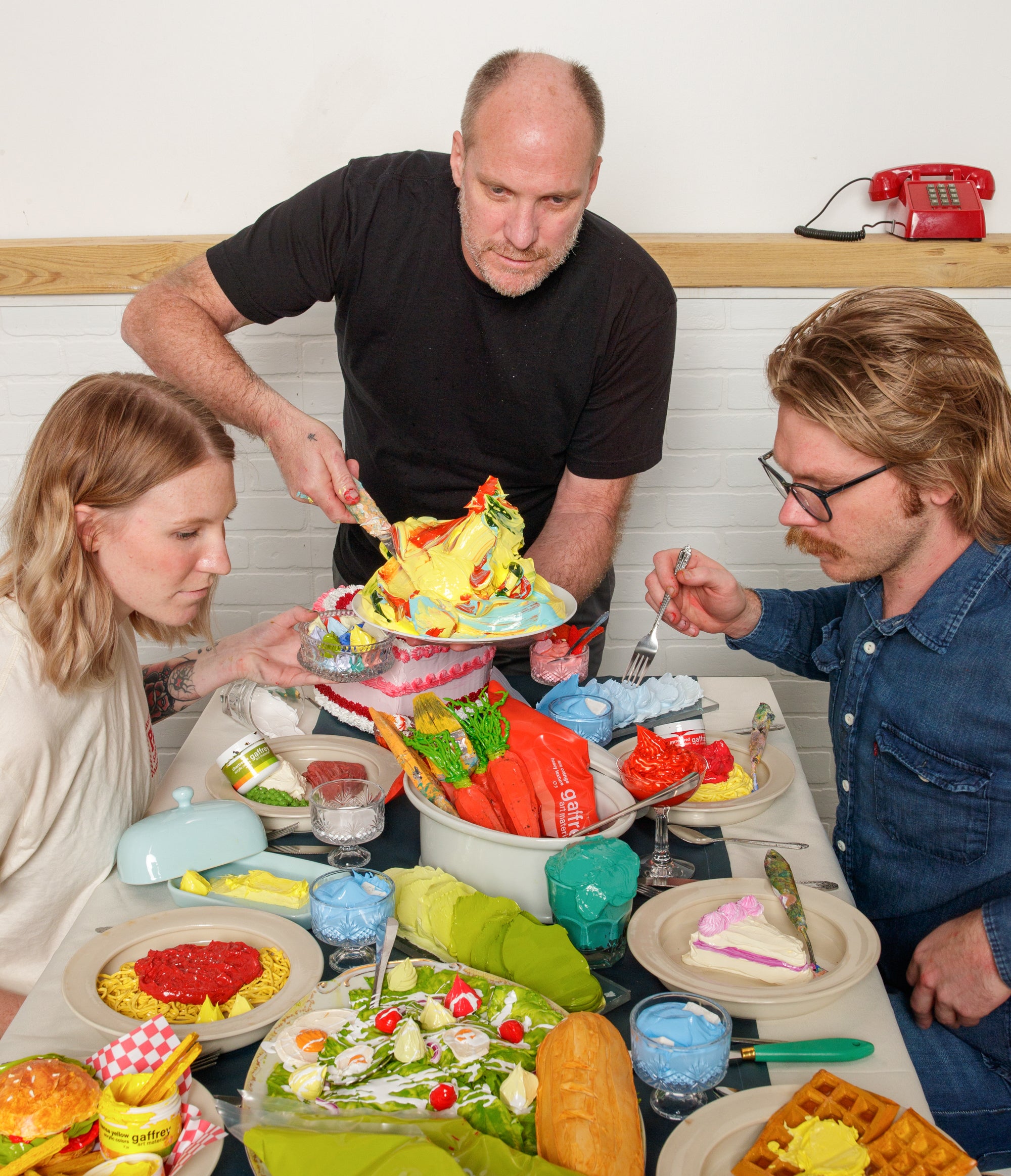 A father serves colorful acrylic paint 'food' sculptures to his adult son and daughter on a long dining table, as part of a whimsical photoshoot by Sean Murphy.