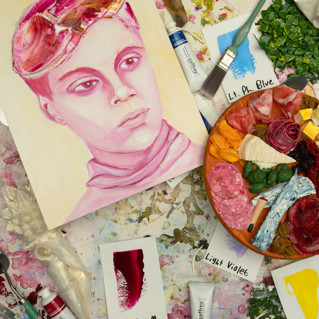 An array of art supplies, acrylic paints, small artworks, and sculptures are scattered across a table