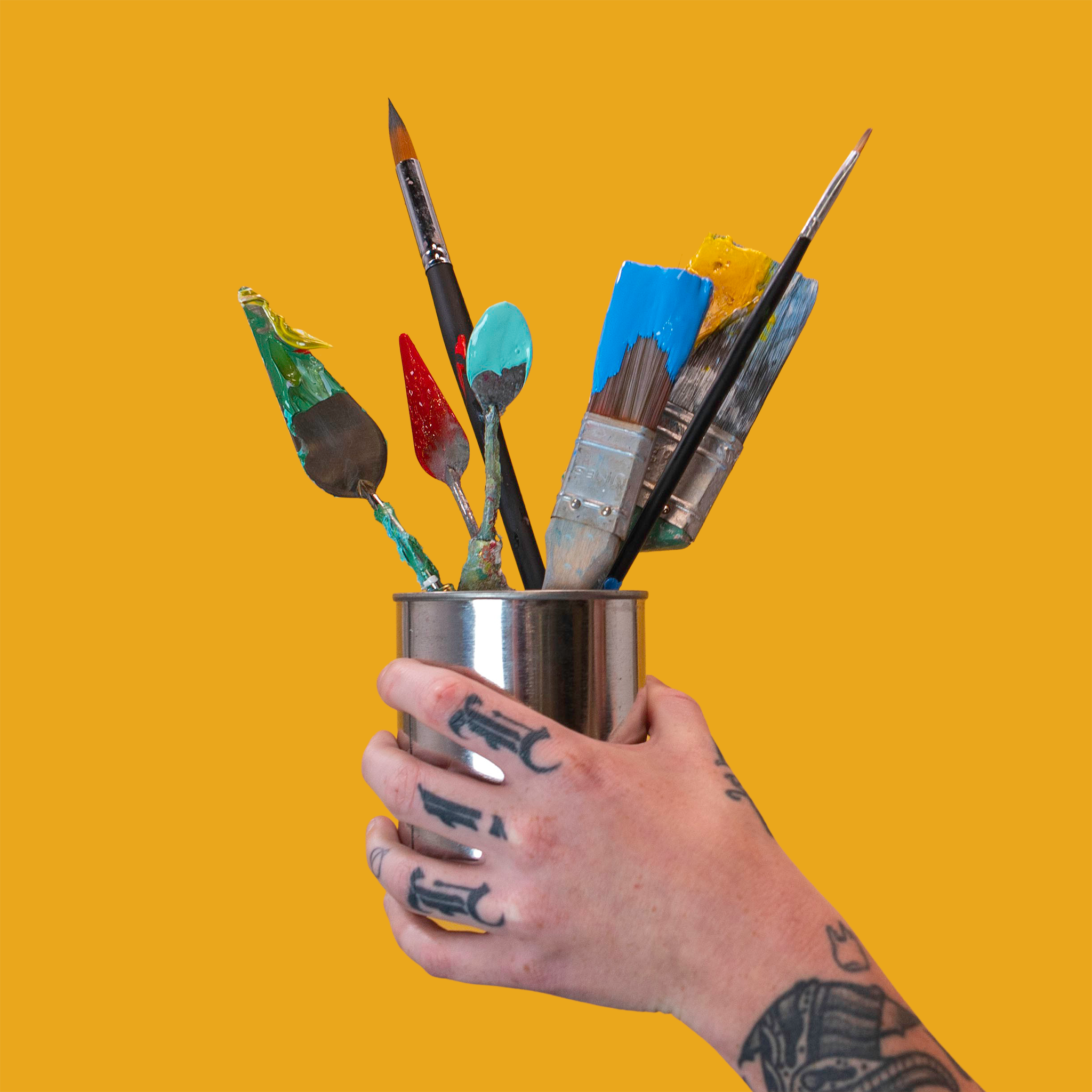 A tattooed artist's hand holds a stainless steel cup filled with various paint-covered tools including brushes and palette knives.